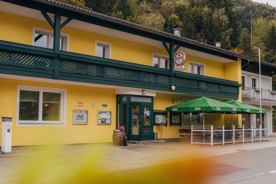 Gelbes Wirtshaus mit gr&uuml;nen Markisen und Schildern, umgeben von B&auml;umen.