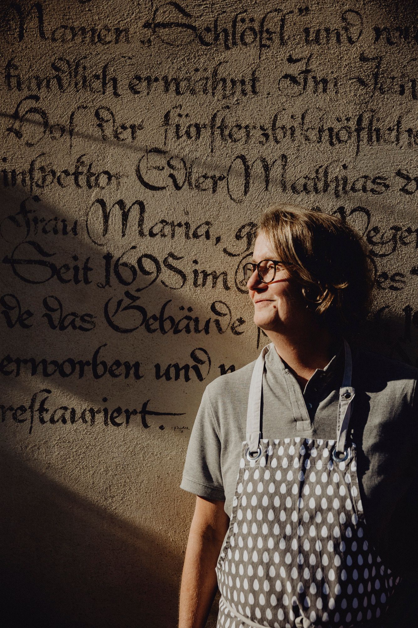 Person mit Schürze vor historischer Wand mit alter Schrift.