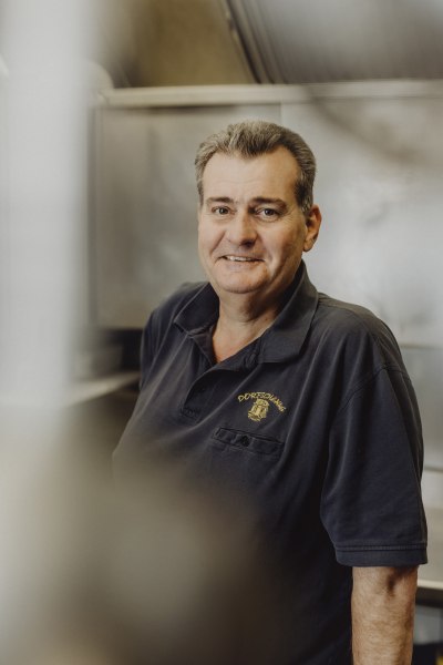 Mann in schwarzem Polohemd mit "Dorfschenke"-Logo l&auml;chelt in die Kamera.