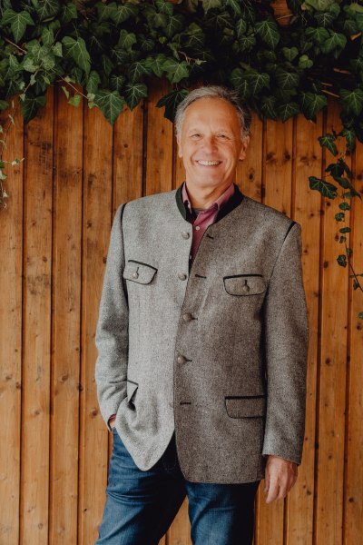 Ein l&auml;chelnder Mann in traditioneller Kleidung vor einer Holzwand mit Efeu.