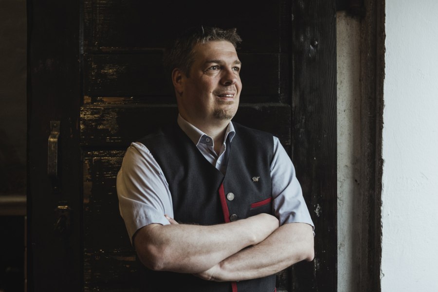 Ein Mann in traditioneller Kleidung steht mit verschr&auml;nkten Armen vor einer dunklen Holzt&uuml;r.