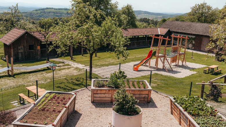 Ein Spielplatz mit Rutsche und Klettergerüst neben Hochbeeten in einem Garten mit Holzhütten im Hintergrund.