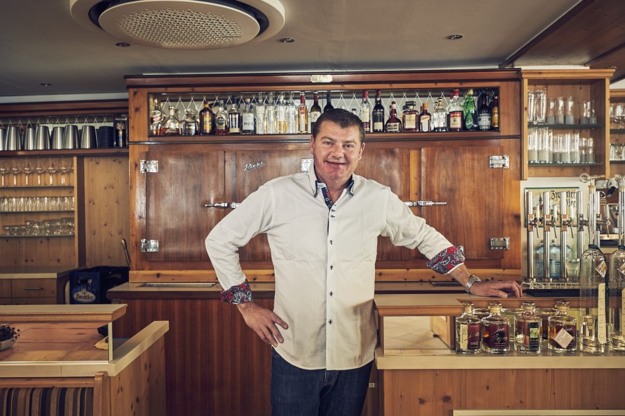 Ein Mann in einem weißen Hemd steht hinter einer Bar mit Flaschen und Gläsern im Hintergrund.