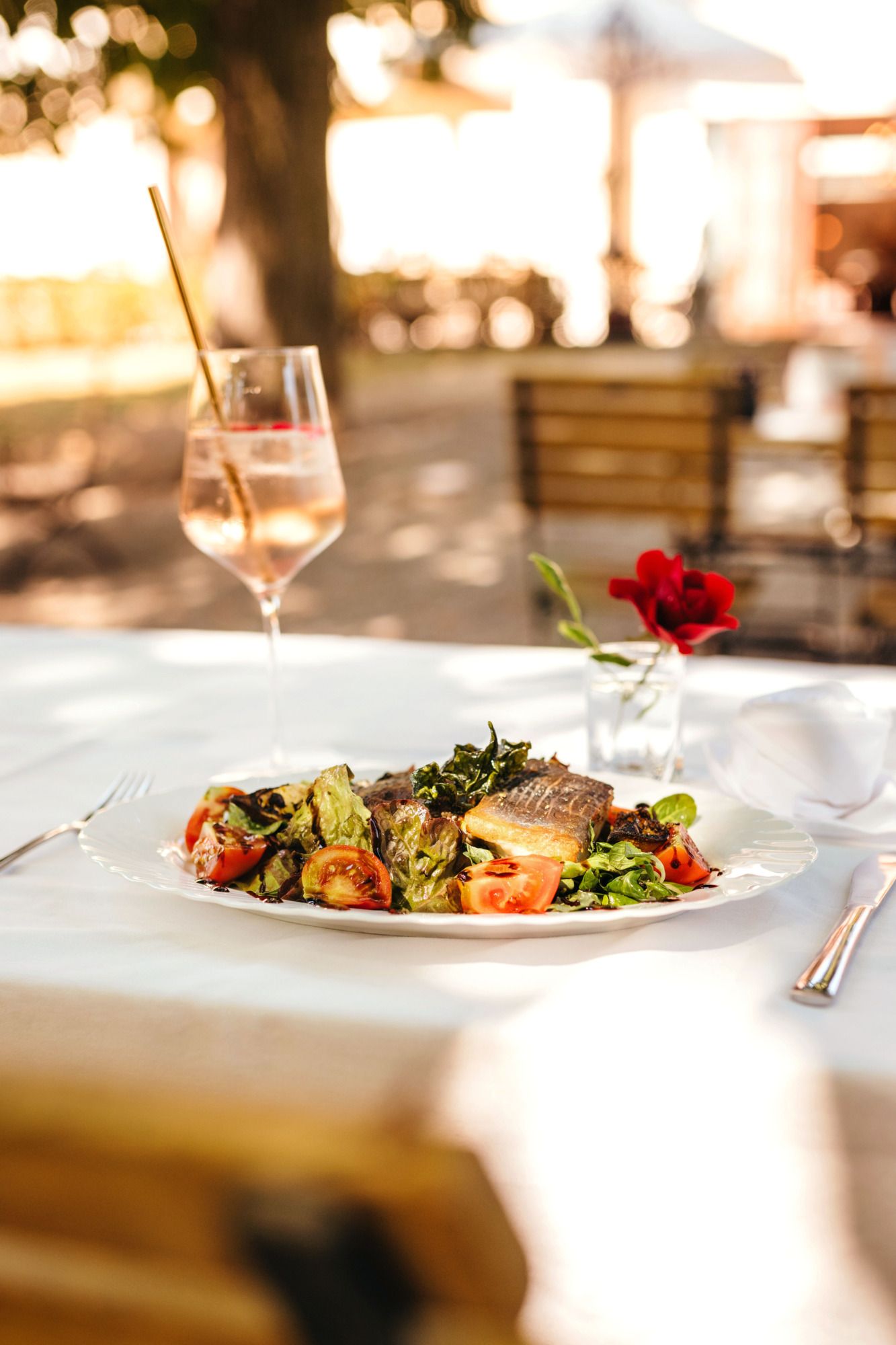 Ein Teller mit Fisch und Salat auf einem Tisch im Freien, daneben ein Glas mit Getränk und eine rote Blume.