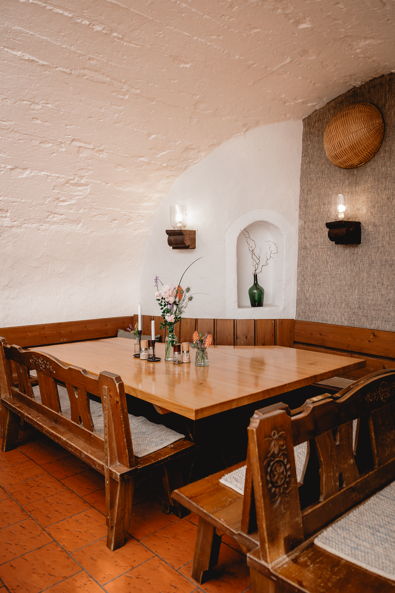 Holztisch mit Eckbänken in einem Raum mit weißem Gewölbe. Auf dem Tisch stehen Blumenvasen, Kerzen und Gewürzstreuer, an der Wand hängen zwei Leuchten.