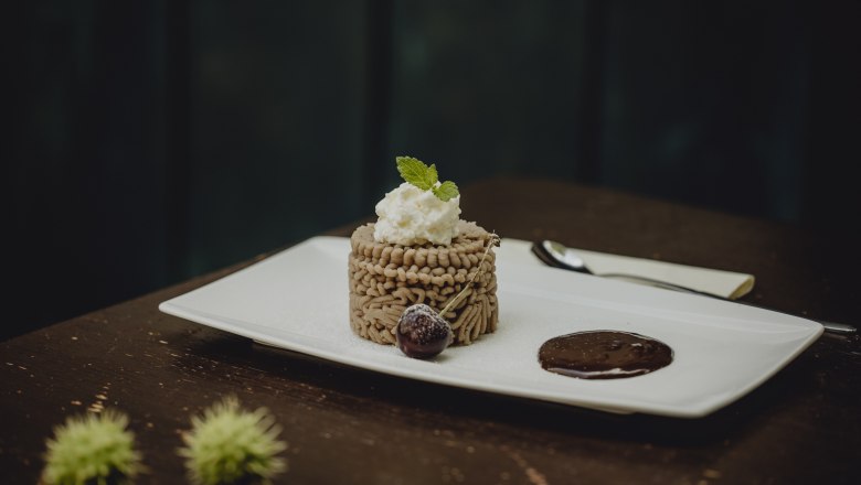 Ein Dessert aus Kastanienmousse auf einem wei&szlig;en Teller, garniert mit Sahne und Minzblatt, daneben eine Schokoladensauce und eine Kirsche.
