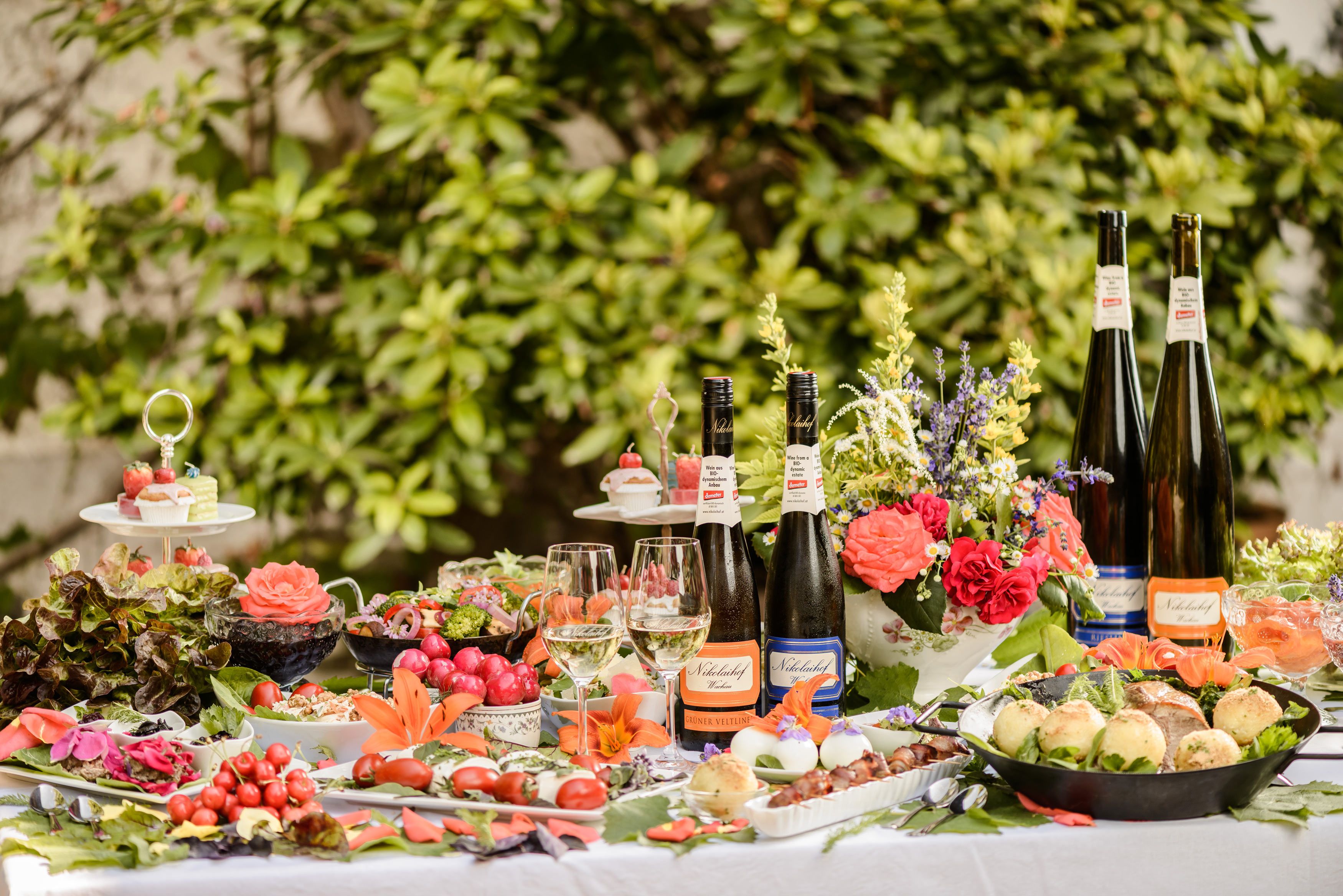 Ein reich gedeckter Tisch im Freien mit Weinflaschen, Gläsern, Blumen und verschiedenen Speisen.