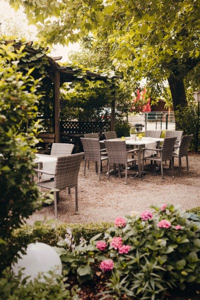 Ein gem&uuml;tlicher Gartenbereich mit Tischen und St&uuml;hlen unter B&auml;umen, umgeben von Pflanzen und Blumen.
