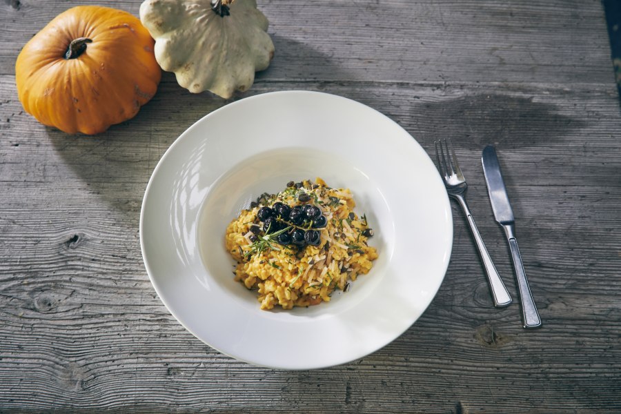 Ein Teller Kürbisrisotto mit Kräutern und Beeren, daneben zwei Kürbisse auf einem Holztisch.