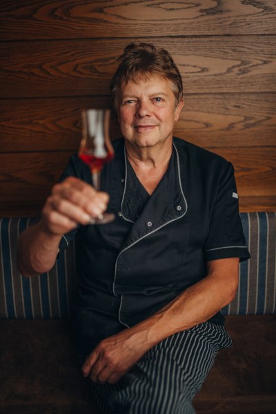Ein Mann in Kochkleidung h&auml;lt ein Glas mit rotem Getr&auml;nk vor einer Holzwand.