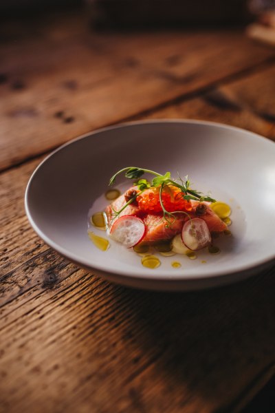 Ein Teller mit gebeizter Lachsforelle, garniert mit Miso, Petersilie, Radieschen und &Ouml;l auf einem Holztisch.