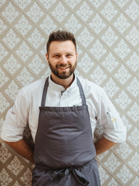 Philipp Essl in Kochkleidung steht vor einer gemusterten Tapete.