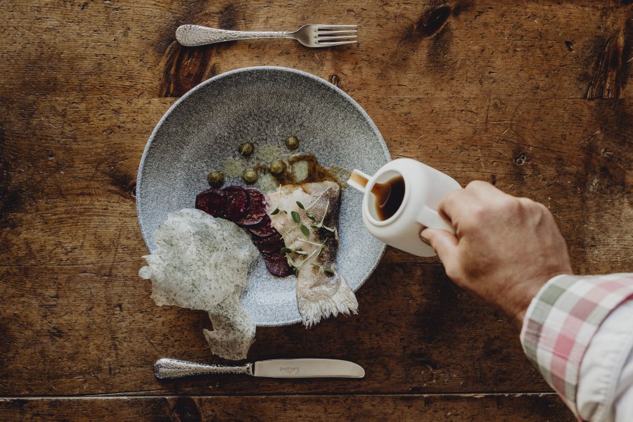 Ein Teller mit Fisch, Roter Bete und Kapern auf einem Holztisch. Eine Hand gie&szlig;t Sauce aus einer Kanne dar&uuml;ber.
