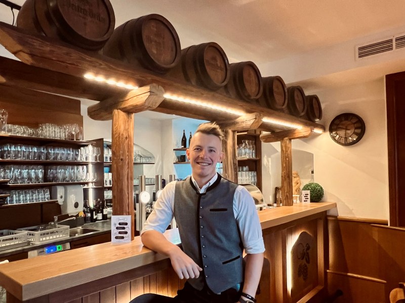 Ein Mann in traditioneller Kleidung steht l&auml;chelnd an einer Bar mit Holzf&auml;ssern und Gl&auml;sern im Hintergrund.