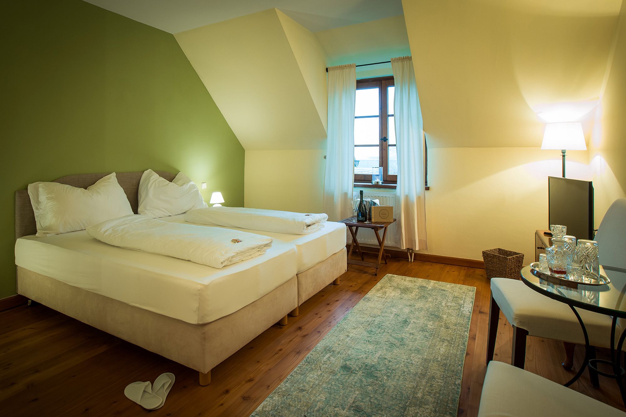 Ein gemütliches Doppelzimmer mit grün gestrichener Wand, zwei Betten, einem Fenster mit Vorhängen, einem kleinen Tisch und einem Fernseher.
