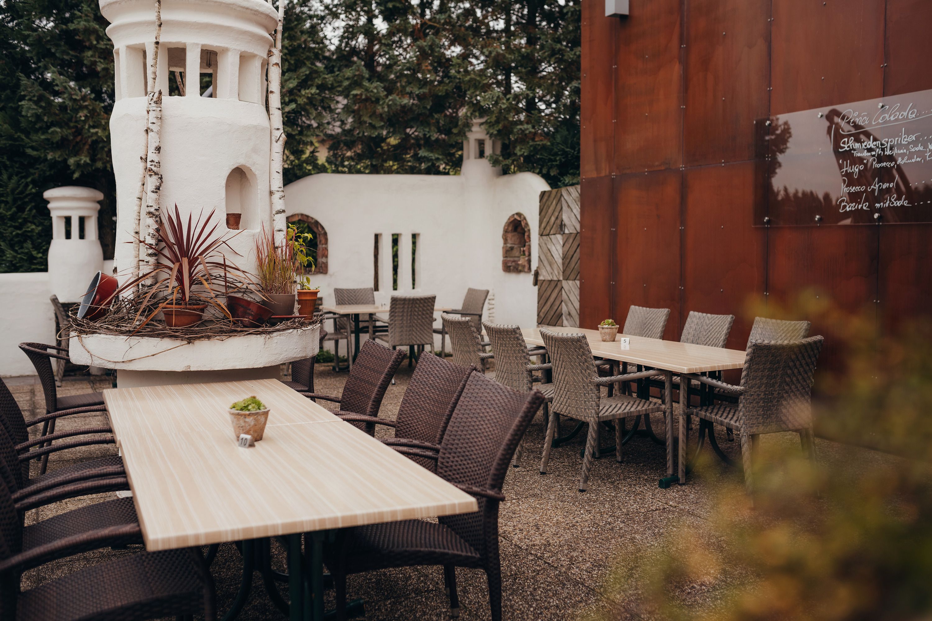 Ein gemütlicher Gastgarten mit Holztischen, geflochtenen Stühlen und dekorativen Pflanzen in einem weißen Turm.