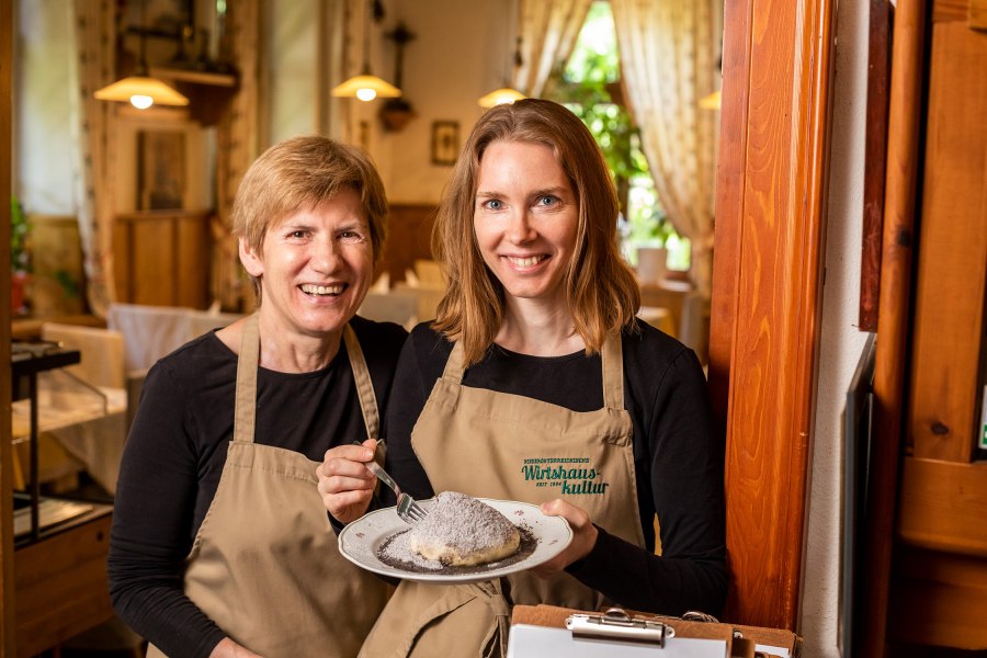 Zwei Frauen in Sch&uuml;rzen halten einen Teller mit einem Dessert in einem gem&uuml;tlichen Restaurant.