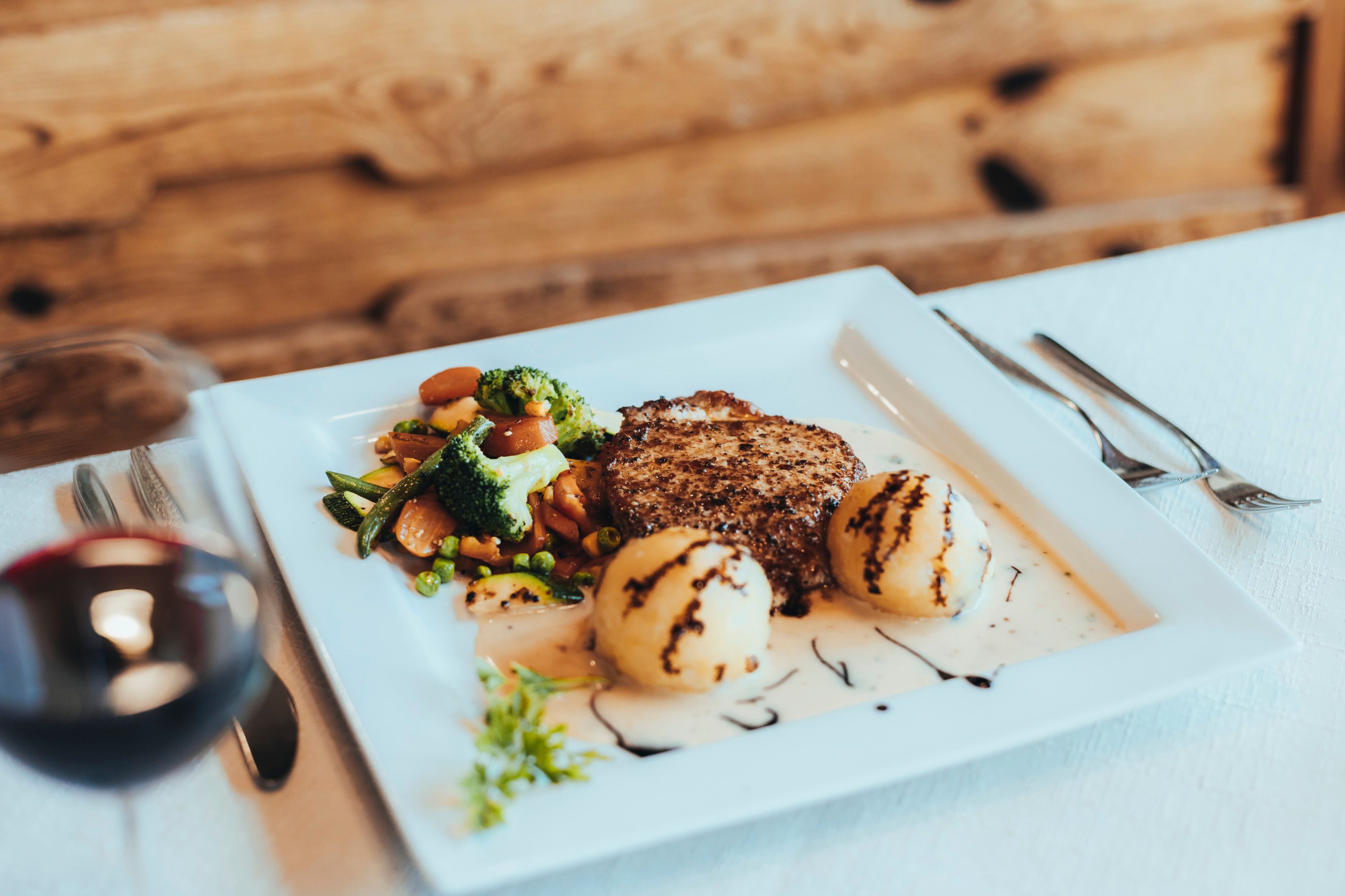 A plate with a chop, vegetables and dumplings, garnished with sauce and balsamic vinegar, on a white table.