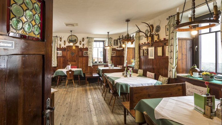 Gemütliche Gaststube mit Holzmöbeln, grünen Vorhängen und Jagdtrophäen an der Wand.