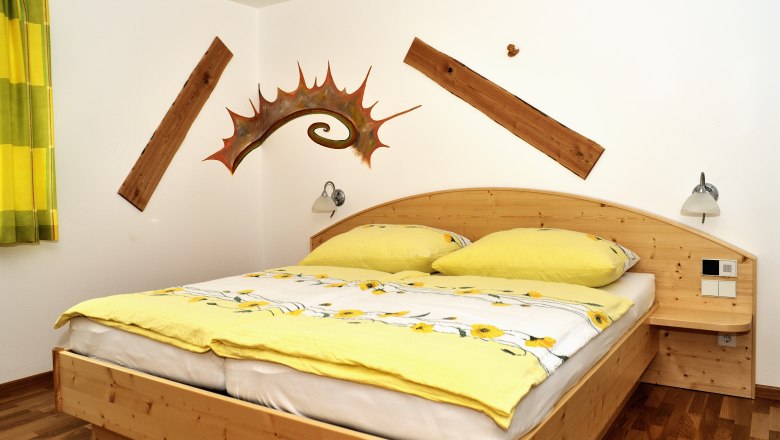 Ein gem&uuml;tliches Schlafzimmer mit einem Holzbett, gelber Bettw&auml;sche und dekorativen Holzelementen an der Wand.