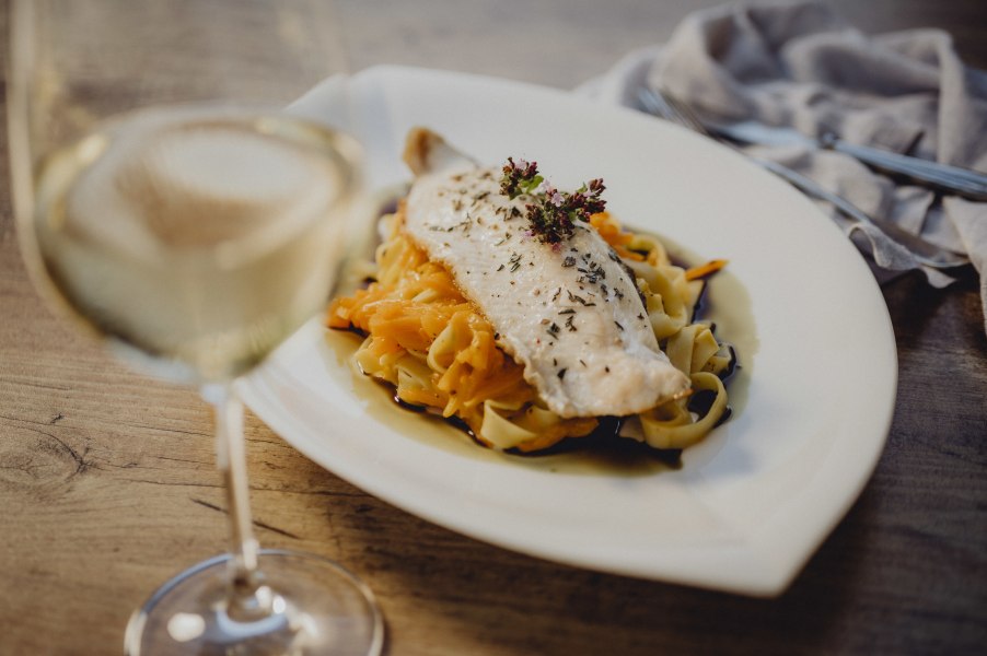 Ein Teller mit Fischfilet auf Nudeln und Gem&uuml;se, daneben ein Glas Wei&szlig;wein.