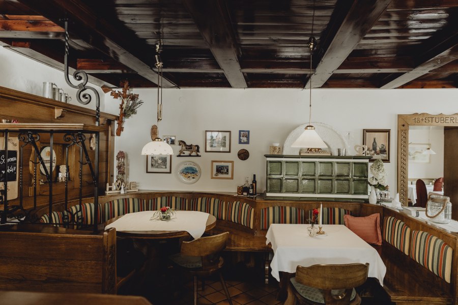 Gem&uuml;tliche Gaststube mit Holzm&ouml;beln, Kachelofen und dekorativen Bildern an der Wand.