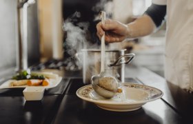 Eine Person schöpft Suppe mit einem Knödel aus einem Topf in einen Teller.