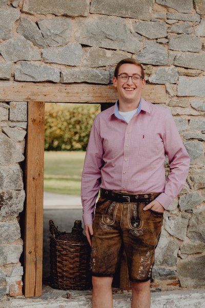 Mann in traditioneller Lederhose und rosa Hemd vor einer Steinmauer.