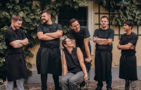 Eine Gruppe von sechs Personen in schwarzer Arbeitskleidung steht und sitzt l&auml;chelnd vor einer mit Weinreben bewachsenen Wand.