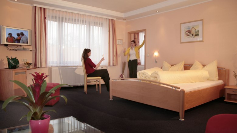 Zwei Frauen in einem Hotelzimmer mit Doppelbett, eine sitzt, die andere steht mit erhobenem Glas.