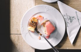 Ein Punschkrapferl auf einem Teller mit einer Gabel, daneben eine Tasse Kaffee und ein Glas Wasser auf einem Tisch.