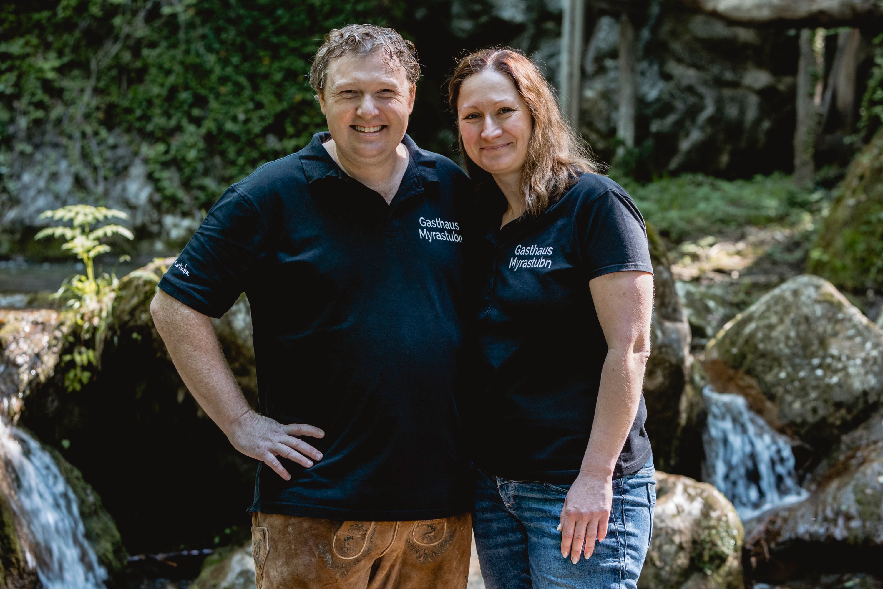 Ein Mann und eine Frau in schwarzen T-Shirts mit der Aufschrift 'Gasthaus Myrastubn' stehen lächelnd vor einem Wasserfall.