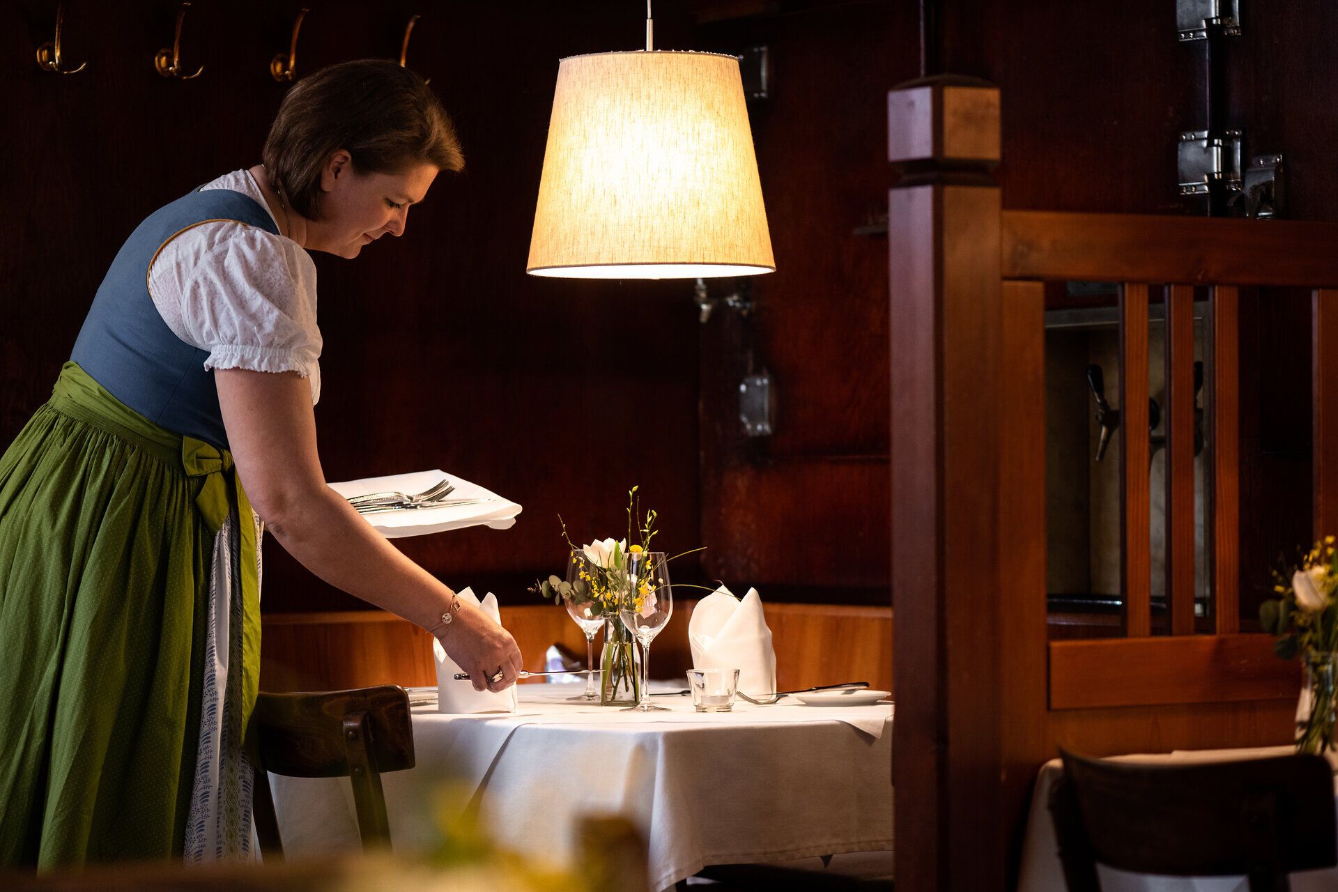 In einem einladenden Wirtshaus wird mit viel Liebe zum Detail ein Tisch für die Gäste gedeckt. Die warmen Holztöne und die sanfte Beleuchtung schaffen eine gemütliche Atmosphäre, die zum Verweilen einlädt. Hier trifft traditionelle Wirtshauskultur auf herzliche Gastfreundschaft.