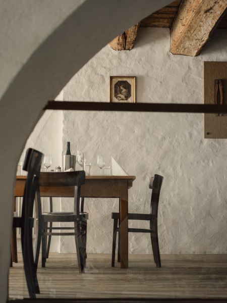 Ein rustikaler Raum mit Holztisch und St&uuml;hlen, dekoriert mit Weinflasche und Gl&auml;sern, unter einer Holzbalkendecke.