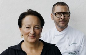 Zwei Personen stehen vor einer weißen Wand. Eine Frau in schwarzer Kleidung und ein Mann in Kochkleidung.