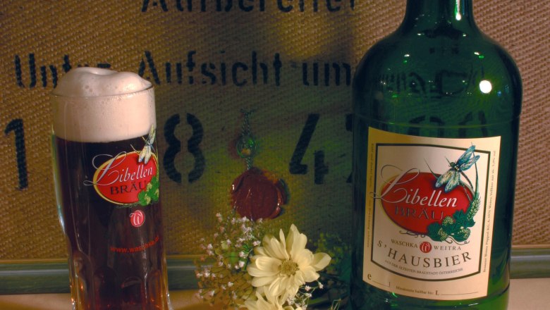 Ein Glas und eine Flasche Libellen Br&auml;u Bier mit Blumen im Vordergrund.
