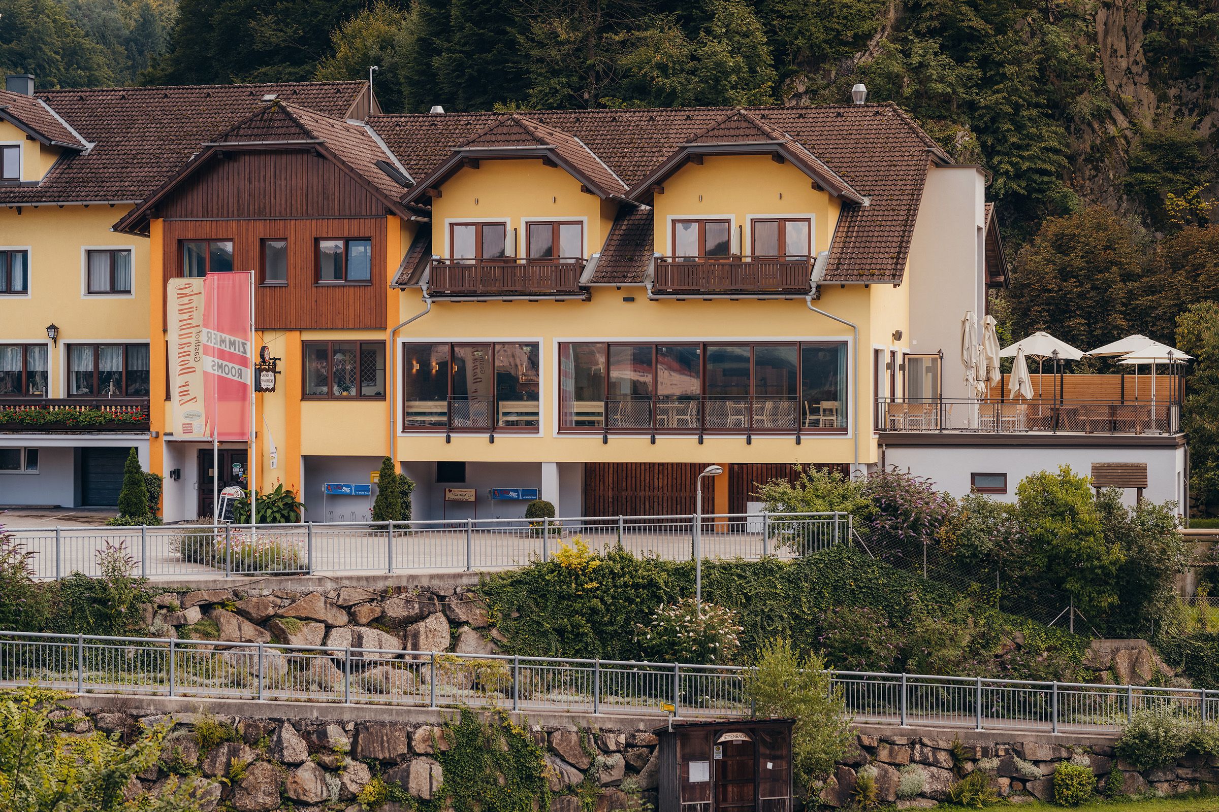 Gelbes Gebäude mit Terrasse und Schildern, umgeben von Bäumen.