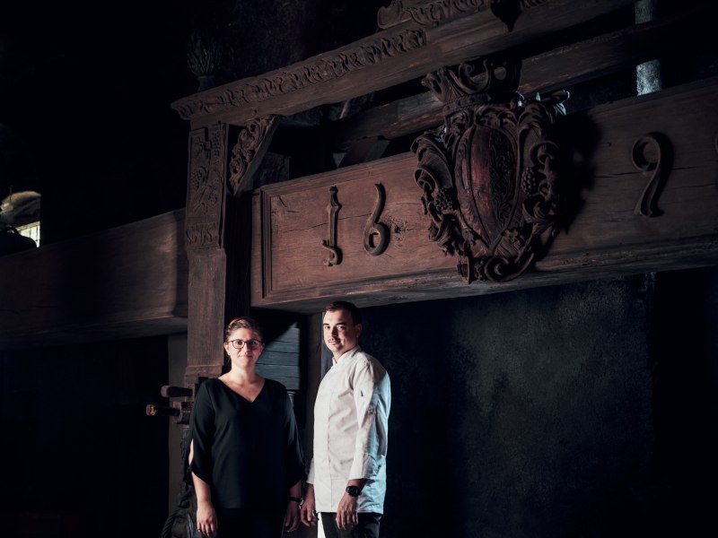 Zwei Personen stehen vor einem alten Holzbalken mit Schnitzereien und der Jahreszahl 1692.