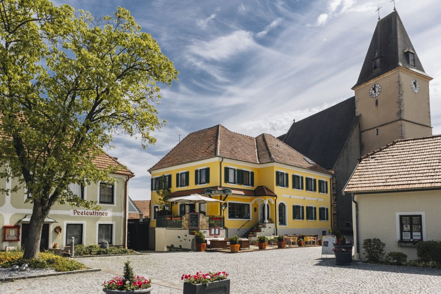 Idyllischer Dorfplatz mit Gasthaus und Terrasse und ein Kirchturm im Hintergrund.