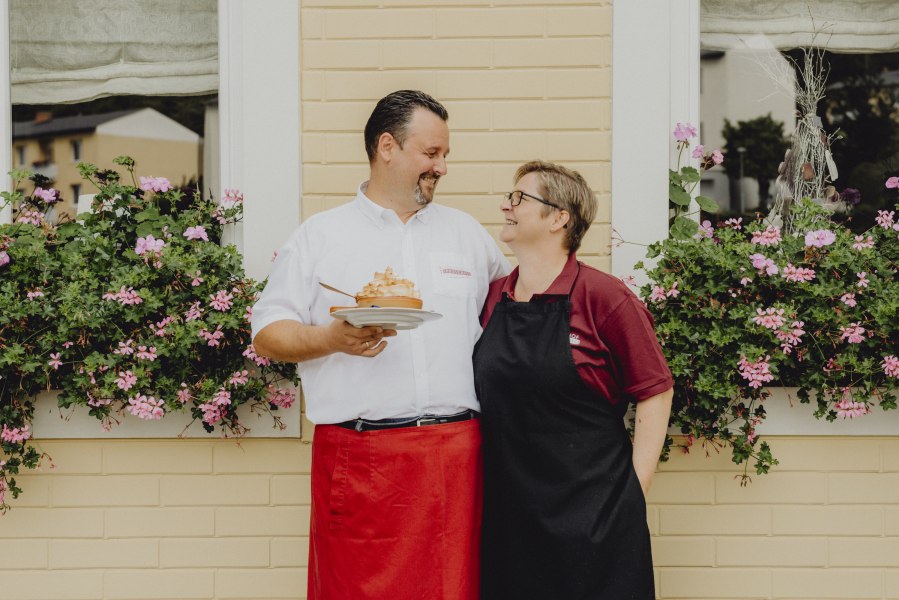 Ein Mann und eine Frau stehen l&auml;chelnd vor einem Fenster mit Blumen.
