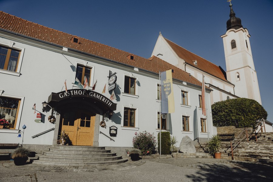 Ein Gasthof mit der Aufschrift 'Gasthof Gamerith' neben einer Kirche mit Turm unter blauem Himmel.