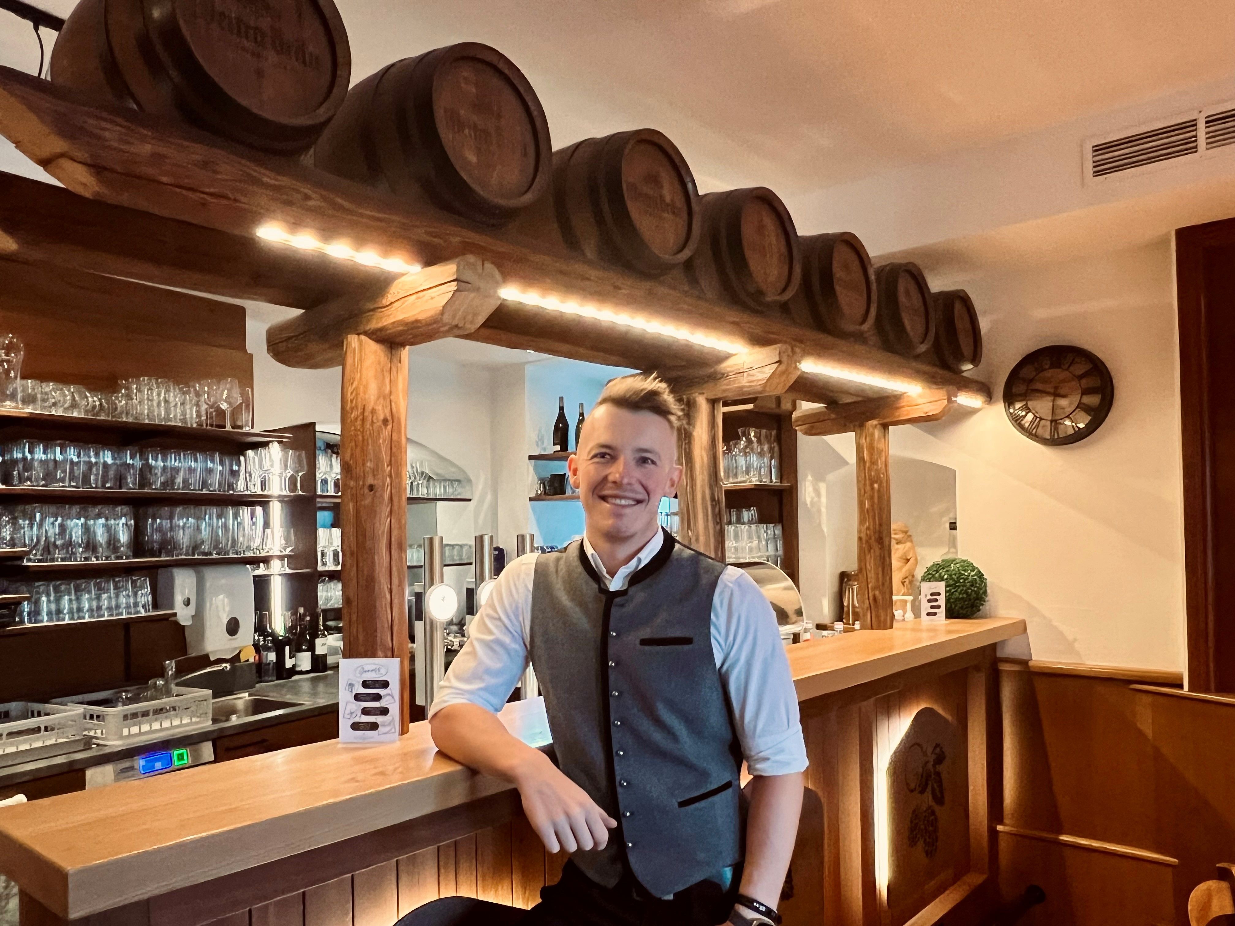 Ein Mann in traditioneller Kleidung steht lächelnd an einer Bar mit Holzfässern und Gläsern im Hintergrund.
