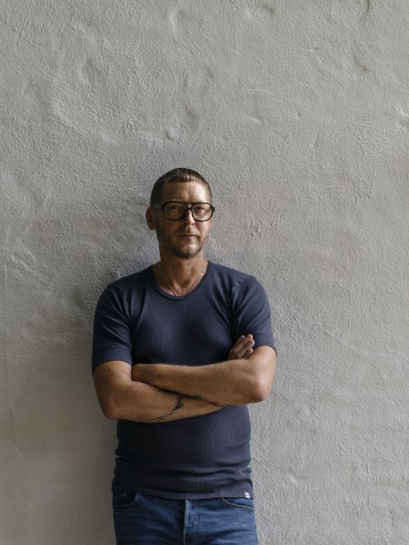 Mann mit Brille und tätowierten Armen steht vor einer weißen Wand.