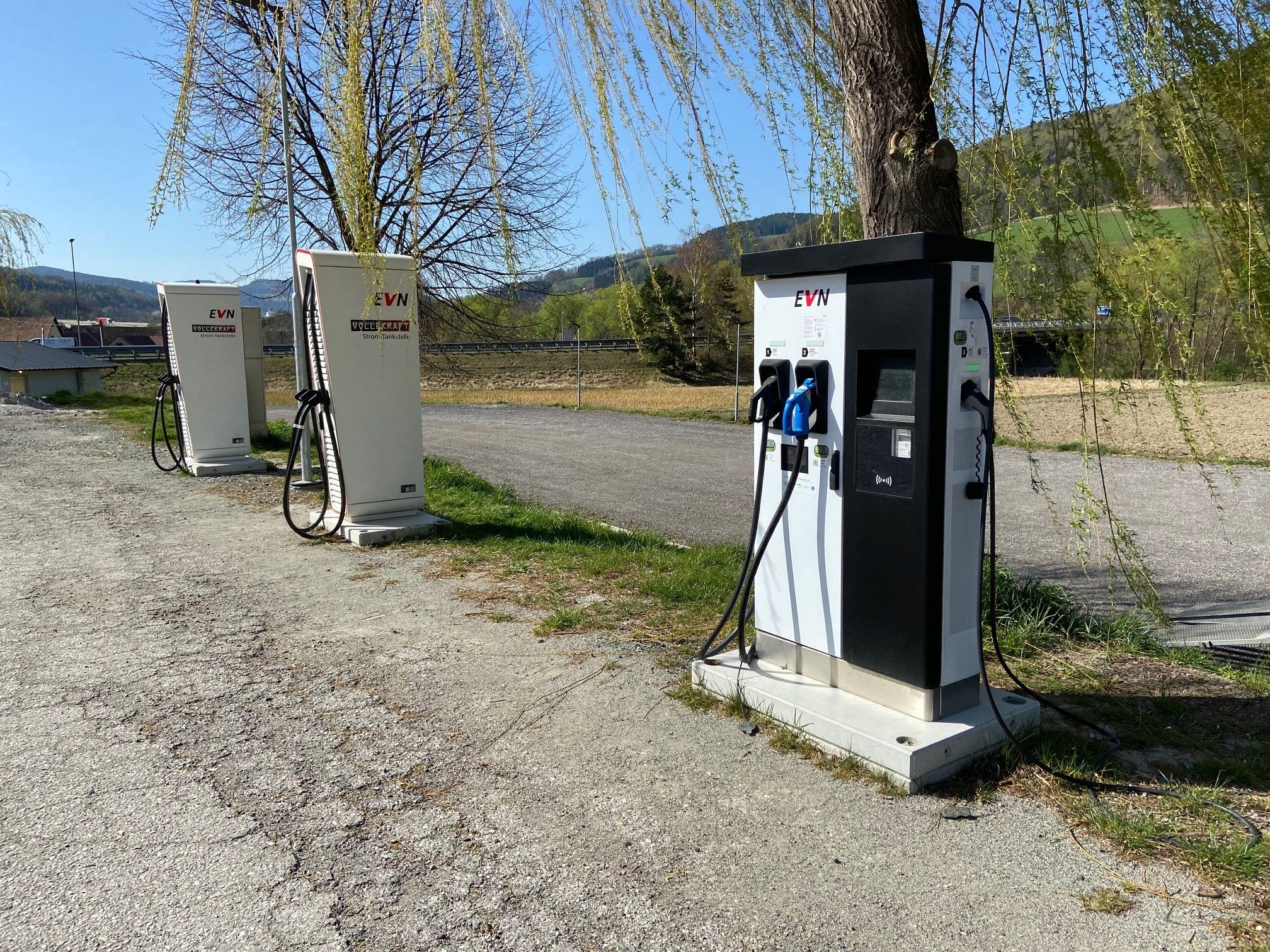 Drei E-Ladestationen auf einem Parkplatz bei einem Gasthof, umgeben von Bäumen und Landschaft.