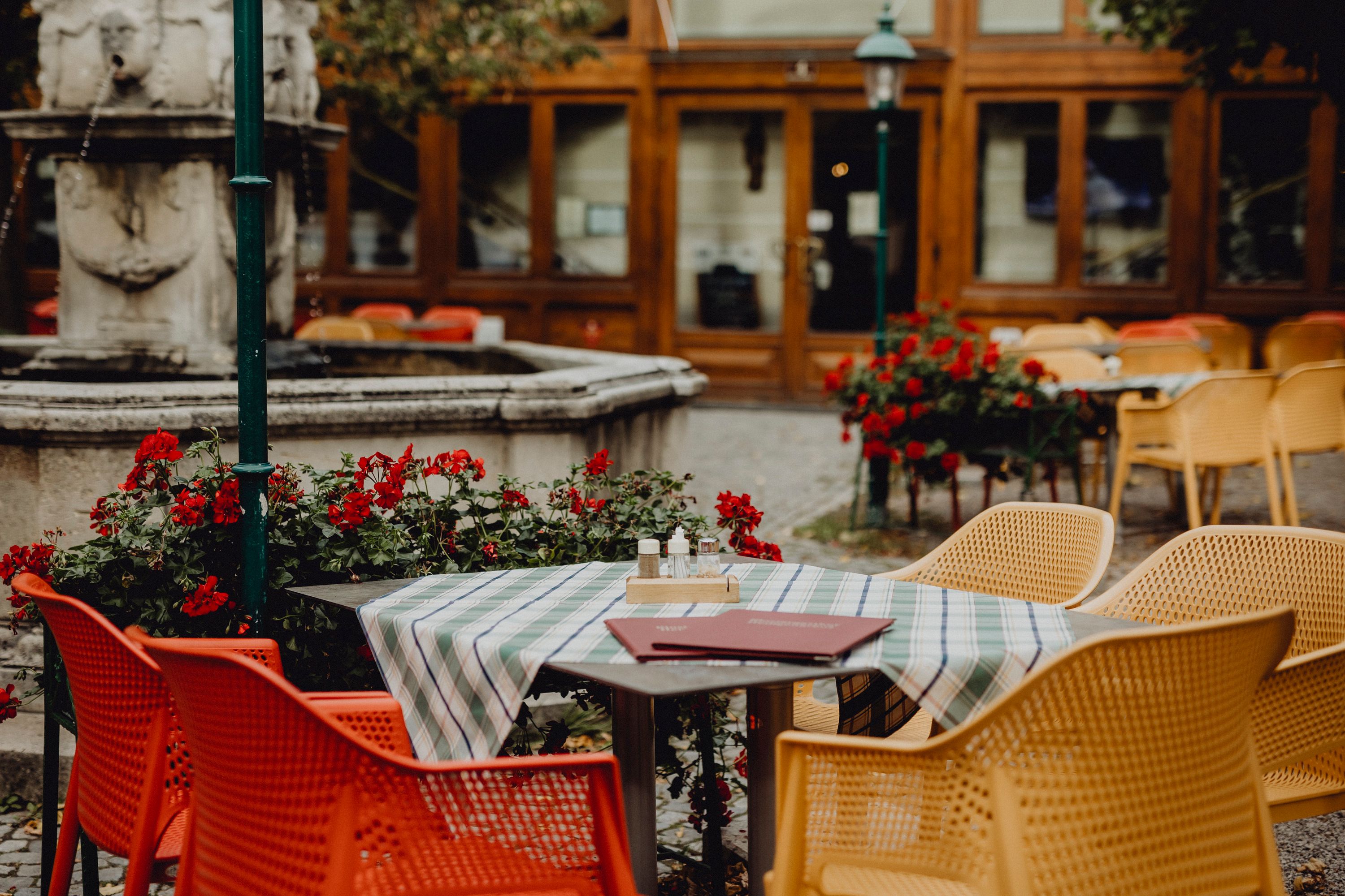Ein Gastgarten mit bunten Stühlen, einem Tisch mit Tischdecke und einem Brunnen im Hintergrund.