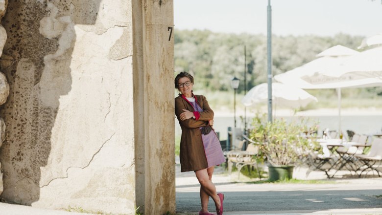 Frau lehnt l&auml;chelnd an einer Mauer, im Hintergrund Terrasse mit Tischen, St&uuml;hlen und Sonnenschirmen.