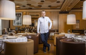 Ein Mann in wei&szlig;er Kochjacke steht in einem eleganten Restaurant mit runden Tischen und gro&szlig;en Lampen.