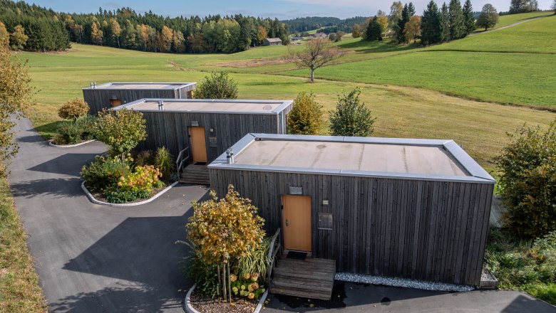 Drei moderne Holzhäuser in einer grünen Landschaft mit Bäumen und Wiesen.
