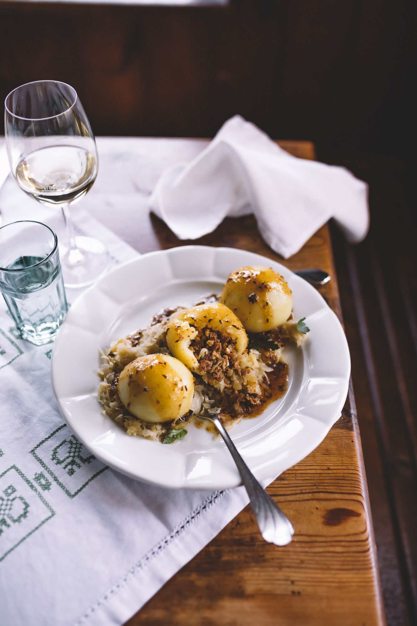 Drei gefüllte Knödel auf Sauerkraut mit Soße auf einem Teller, daneben ein Glas Weißwein.