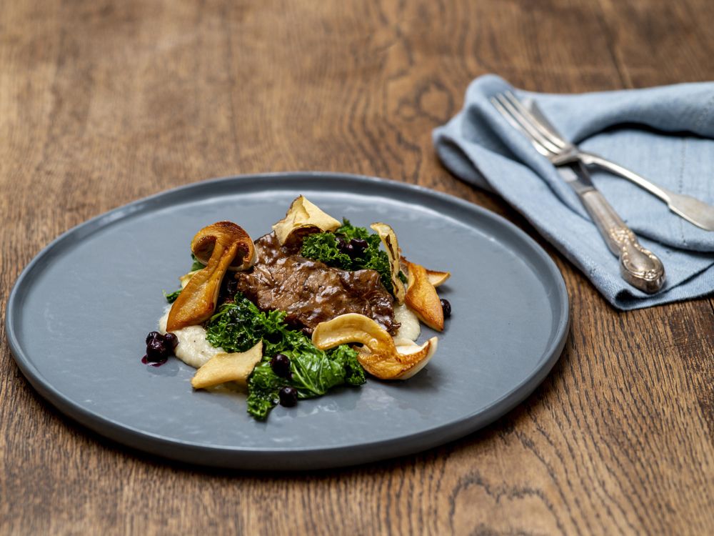 Gourmetgericht mit Fleisch, Pilzen und Grünkohl auf grauem Teller.